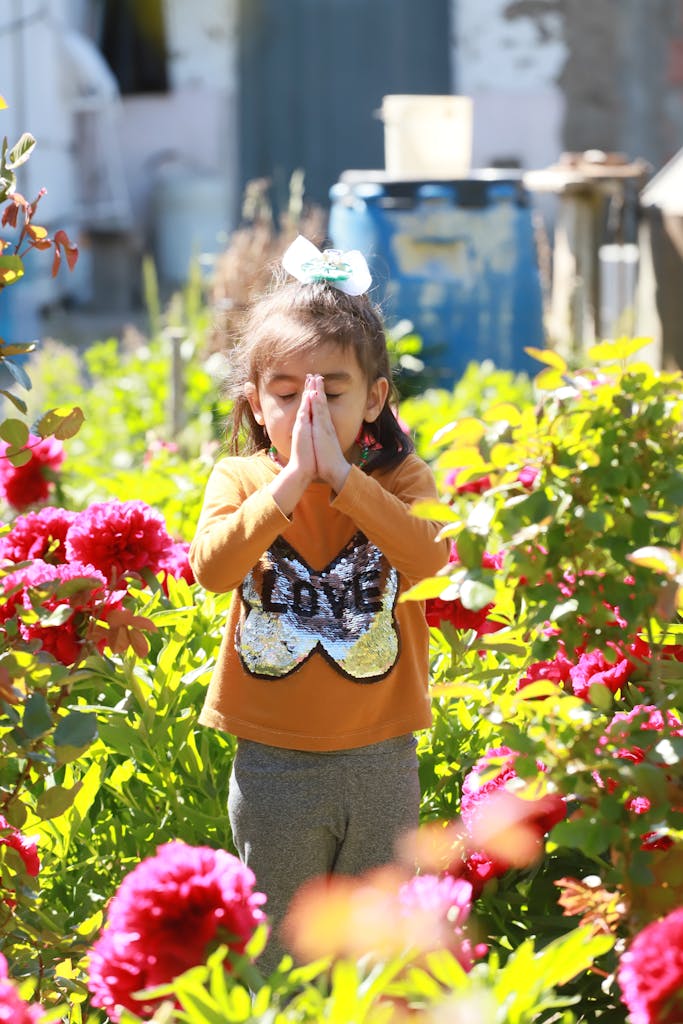 A child stands praying with closed eyes in a colorful rural garden filled with flowers.