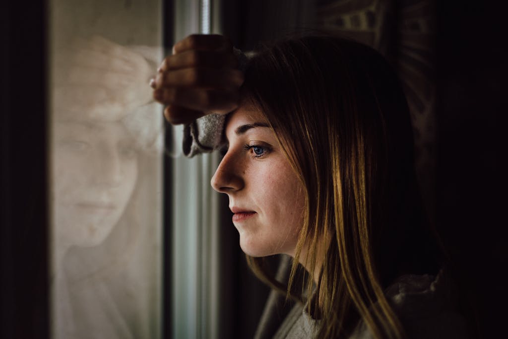 A contemplative young woman looks out a window with a reflective expression, indoors.