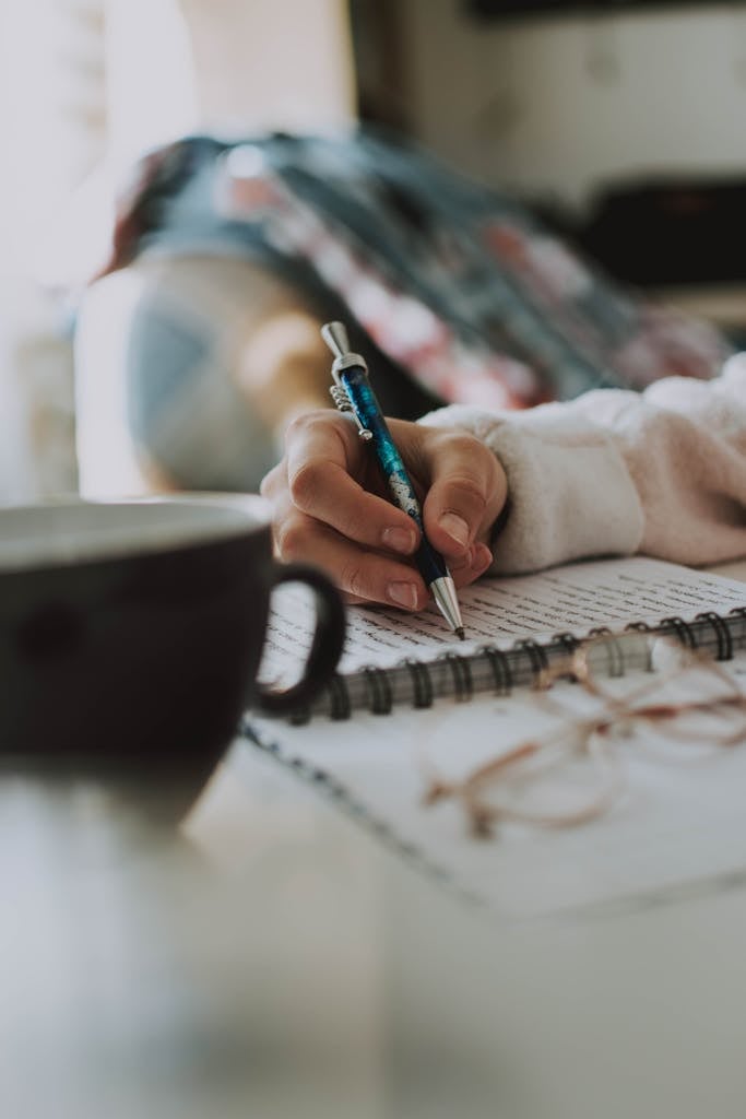 A person writes in a notebook while a cup of coffee sits nearby, indoors.