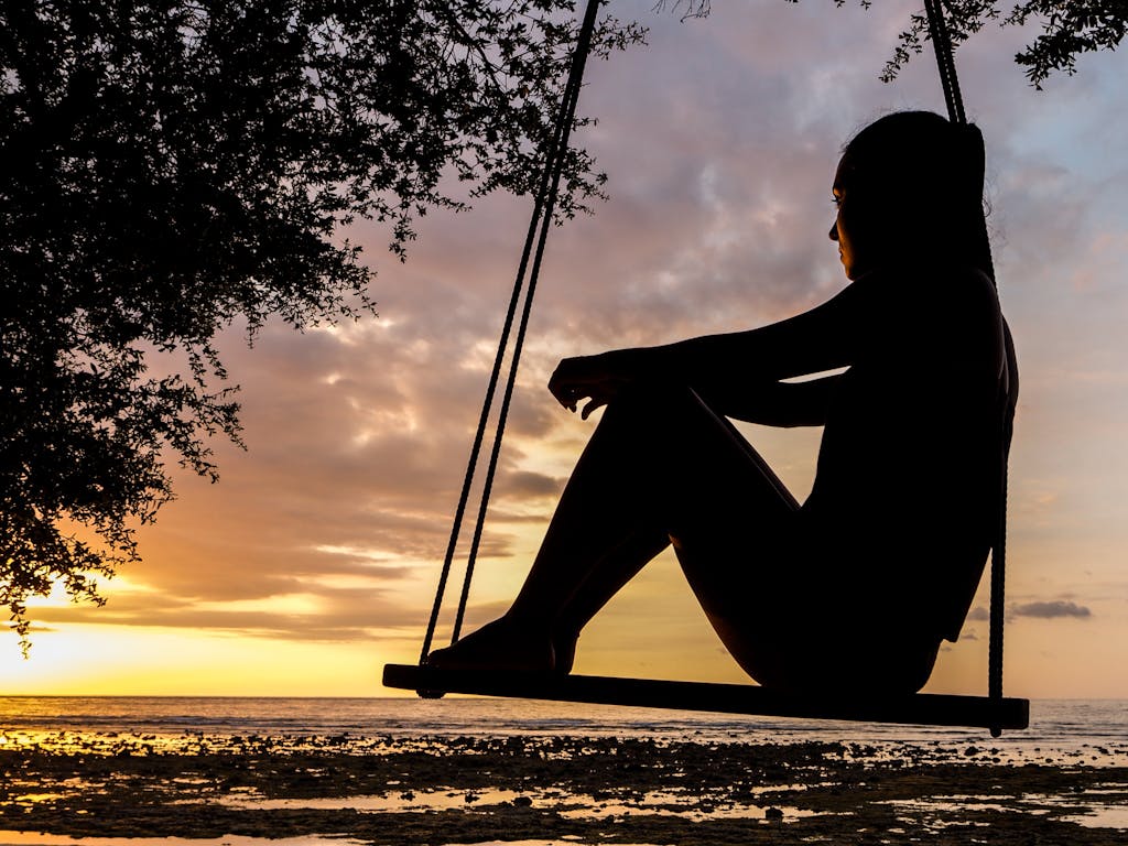 A serene sunset silhouette of a woman sitting on a swing by the seashore, evoking tranquility and reflection.