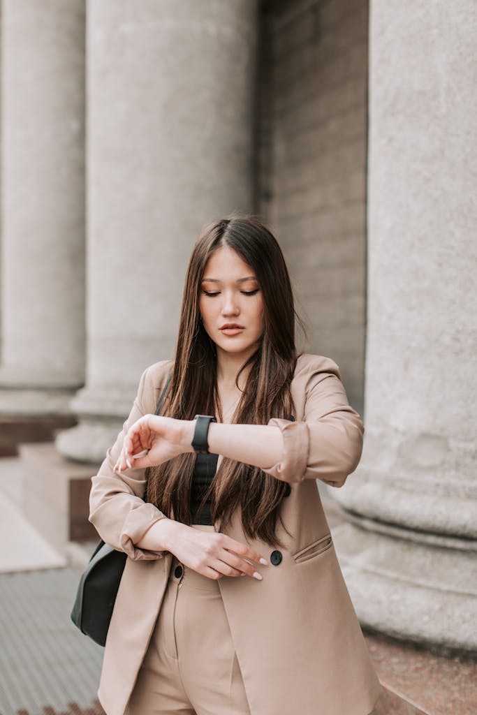 Asian businesswoman in a beige blazer checking time on wristwatch outside a building with columns.