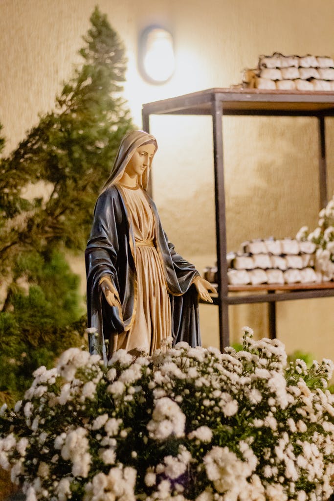 Statue of Virgin Mary surrounded by white flowers, indoors with warm lighting.